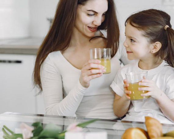 Happy mother and daughter in casual clothes drinking juice and looking at each other while having breakfast together in contemporary kitchen
